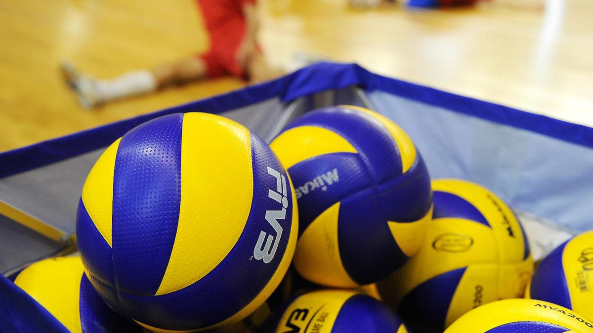 Stos piłek do siatk&oacute;wki w koszu, na pierwszym planie widoczne są piłki niebiesko-ż&oacute;łte. W tle zamazane sylwetki os&oacute;b w czerwonych i niebieskich strojach na hali sportowej.