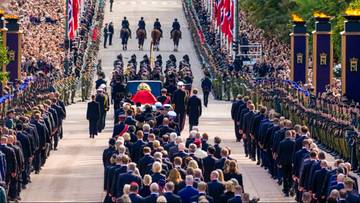 Uroczysta procesja żałobna z trumną niesioną ulicą otoczoną szpalerem wojska i tłumem ludzi.