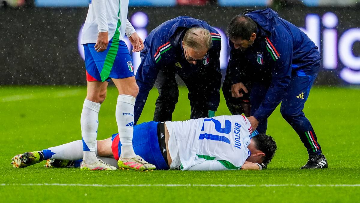 Piłkarz reprezentacji w białej koszulce z niebieskimi elementami leży na trawie, przytrzymując się ręką za twarz. Dookoła niego stoją członkowie sztabu medycznego w niebieskich kurtkach, udzielając pomocy.