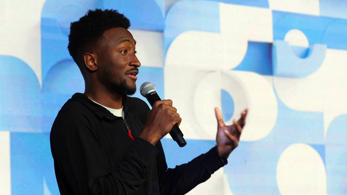 Mężczyzna z mikrofonem w ręku, przemawiający na scenie na tle niebiesko-białej grafiki.