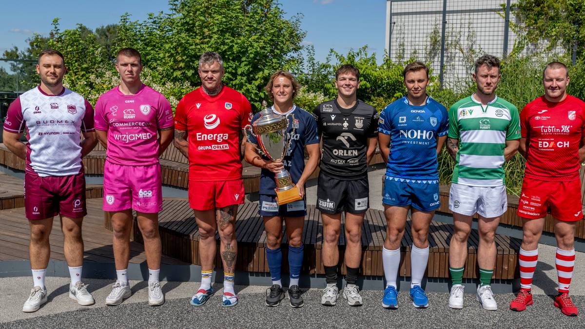 Grupa ośmiu mężczyzn, zawodników rugby, pozuje do zdjęcia na zewnątrz. W środku grupy jeden z zawodników trzyma w rękach puchar.