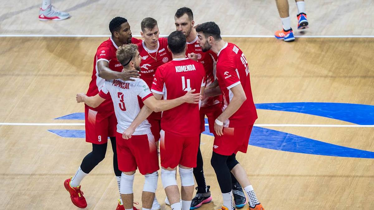 Grupa polskich siatkarzy w czerwonych strojach z białymi numerami stoi w okręgu i trzyma się za ramiona, okazując radość po meczu.