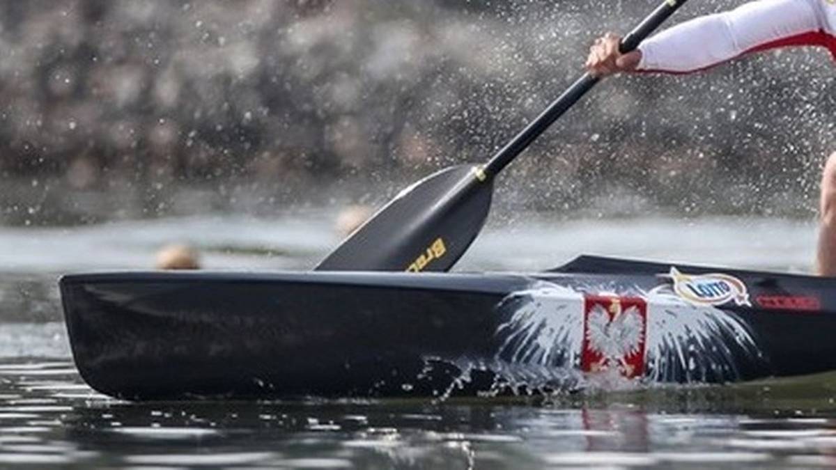 Dłoń osoby trzymająca wiosło, wiosłująca w czarnej łodzi kajakowej z biało-czerwonym godłem Polski na burcie. Woda rozpryskuje się wokół łodzi.