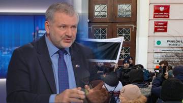 Waldemar Żurek w studiu telewizyjnym obok kolażu przedstawiającego budynek z tablicami informacyjnymi i grupę ludzi.