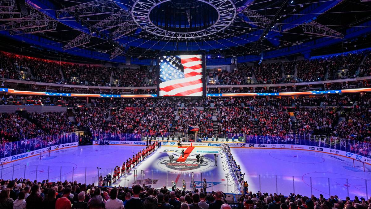 Widok na pełną trybun kibiców w hali sportowej podczas meczu hokejowego, z lodowiskiem i wyświetlaną na ekranie amerykańską flagą.