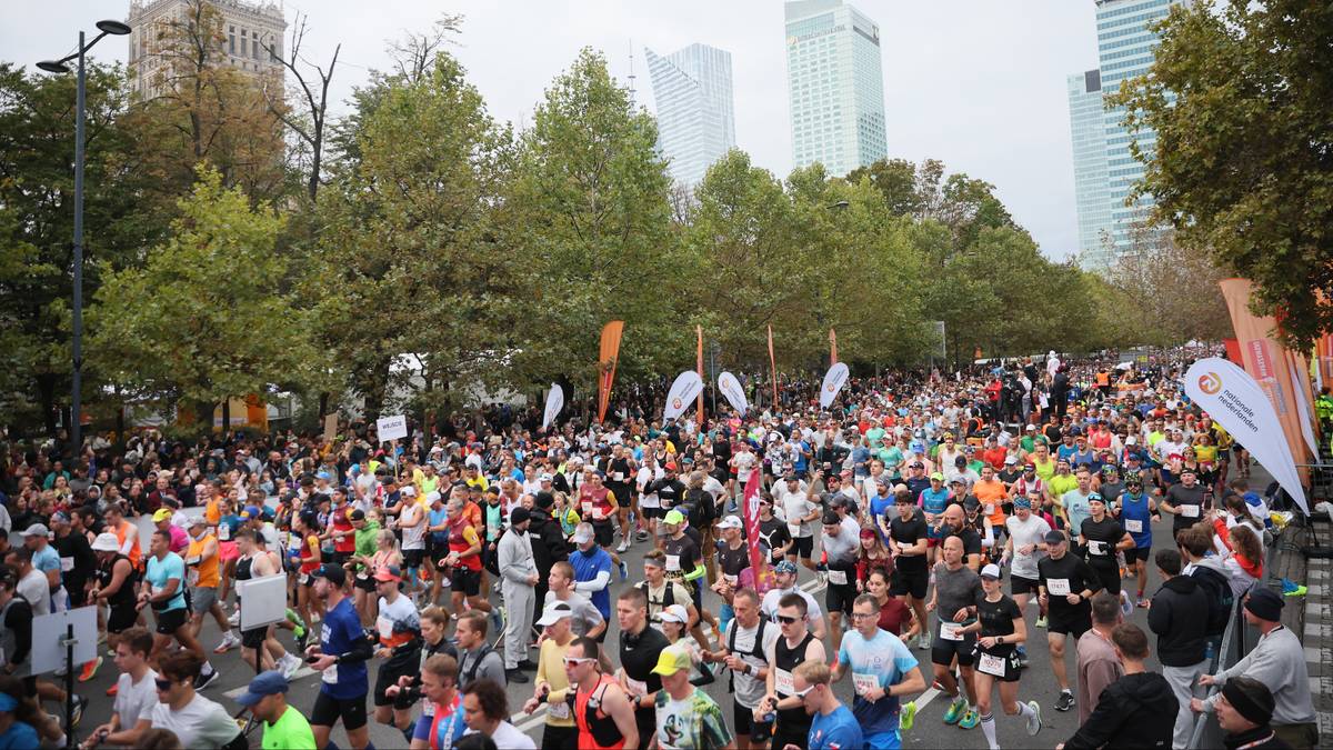 Duża grupa biegaczy zebrana na starcie zawod&oacute;w sportowych, z miejskimi budynkami w tle.