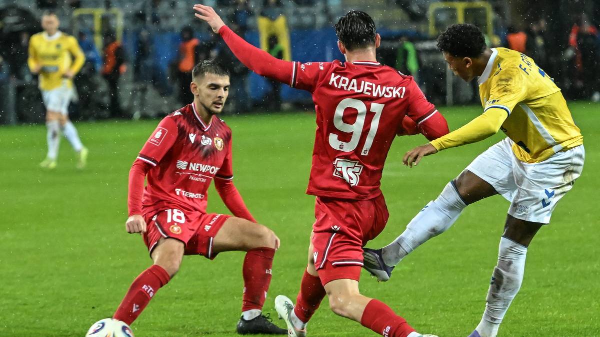 Piłkarz w czerwonym stroju z numerem 91 na plecach próbuje odebrać piłkę przeciwnikowi w żółtej koszulce. Obok nich znajduje się drugi zawodnik w czerwonym stroju.