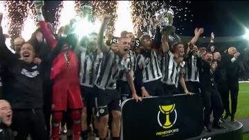 Drużyna piłkarska świętująca zdobycie trofeum, z fajerwerkami w tle.