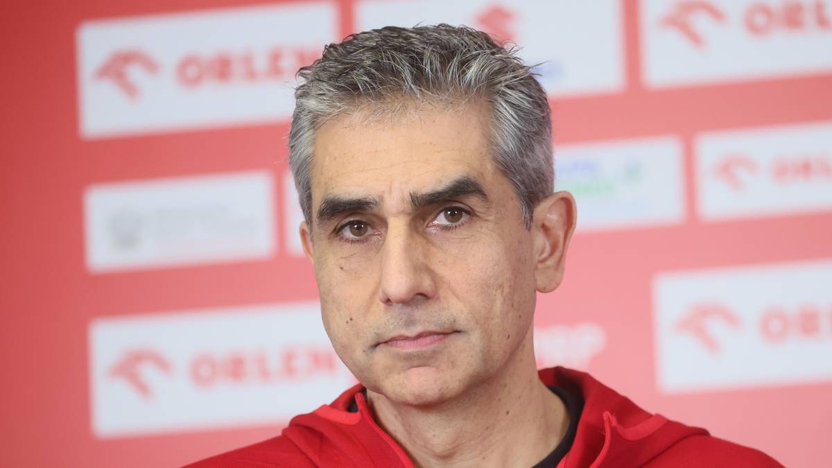 Portret mężczyzny w średnim wieku, ubranego w czerwoną bluzę sportową z herbem Polski, na tle ścianki z logo sponsora.