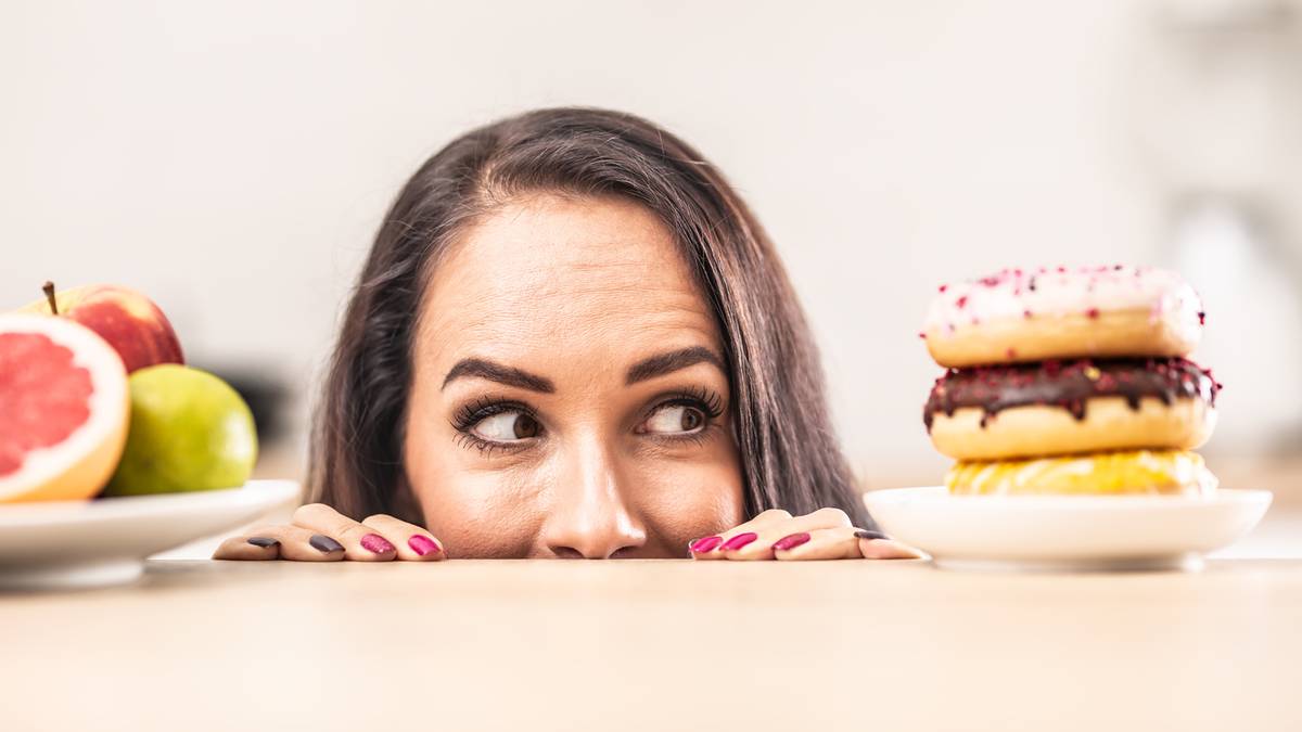 Kobieta z zaciekawieniem spogląda na stos pączków obok talerza z owocami.