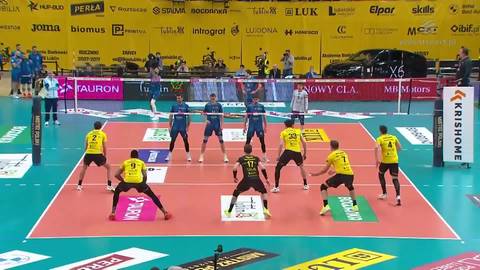 Siedmiu zawodnik&oacute;w w ż&oacute;łtych strojach i jeden zawodnik w niebieskim stroju grają w siatk&oacute;wkę na boisku. Na bandach otaczających boisko widnieją loga sponsor&oacute;w.