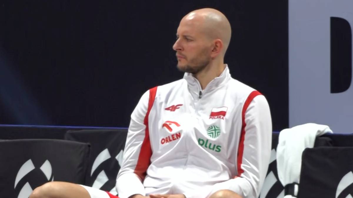 Siatkarz Bartosz Kurek siedzi na ławce, ubrany w białą bluzę z czerwonymi elementami i logotypami, wpatrując się w bok.