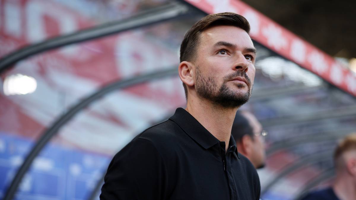 Portret mężczyzny w czarnej koszulce polo, z kr&oacute;tką brodą, patrzącego w bok i w g&oacute;rę. W tle widać fragment stadionu.