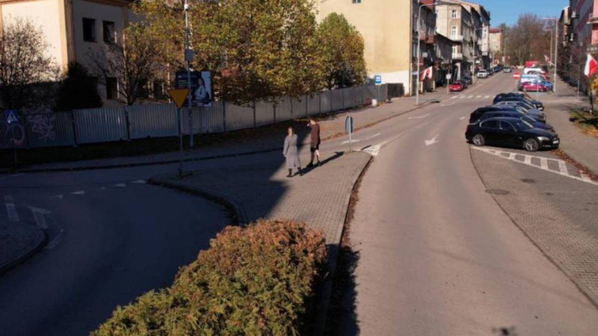 Widok na miejską ulicę z chodnikiem, budynkami i zaparkowanymi samochodami.