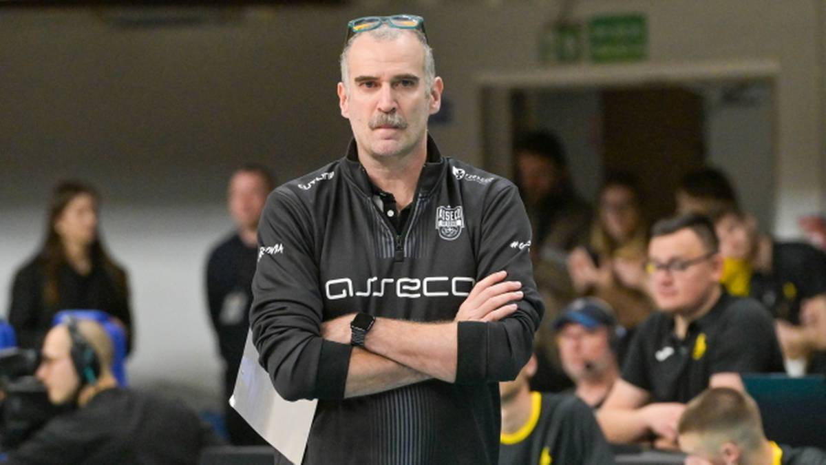 Trener w czarnej kurtce z napisem "Asseco" stoi ze skrzyżowanymi rękami.