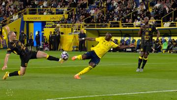 Dw&oacute;ch piłkarzy w akcji na boisku. Jeden w ż&oacute;łtej koszulce i spodenkach pr&oacute;buje kopnąć piłkę, drugi w czarnej koszulce i spodenkach stara się mu przeszkodzić.