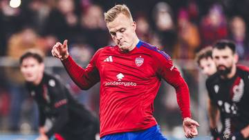 Piłkarz w czerwonej koszulce i niebieskich spodenkach kopie piłkę na zielonym boisku.