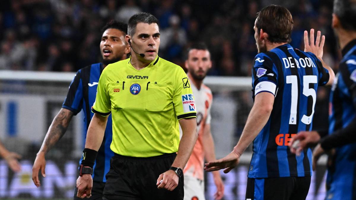 Sędzia piłkarski w żółtej koszulce i czarnych spodenkach stoi na boisku, otoczony przez zawodników w niebiesko-czarnych strojach.