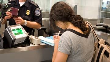 Funkcjonariusz Straży Granicznej USA w mundurze sprawdza paszport w ręku kobiety przy stanowisku kontrolnym.