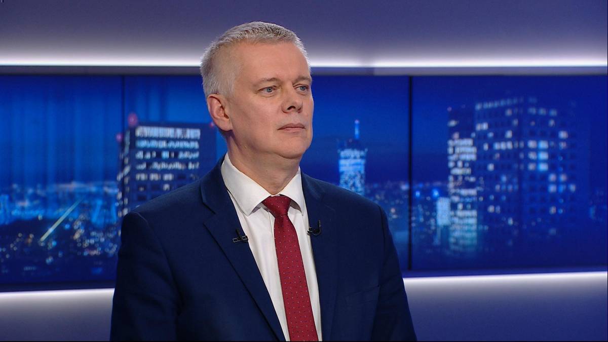 Mężczyzna w garniturze i krawacie w studiu telewizyjnym na tle niebieskiej scenografii.