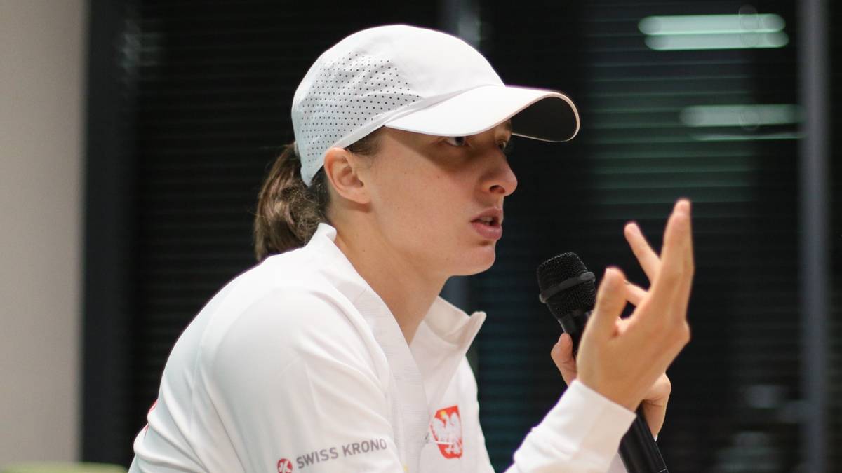 Iga Świątek przemawia do mikrofonu, siedząc przy stole w białej czapce i bluzie.