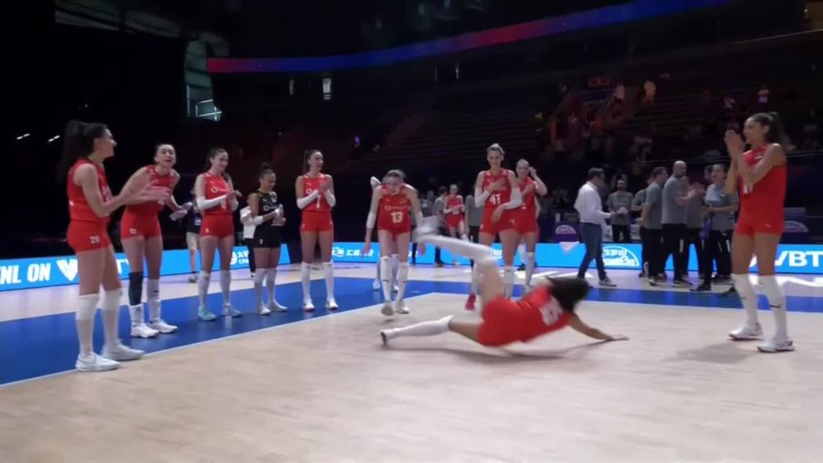 Grupa zawodniczek siatkówki w czerwonych strojach stoi po jednej stronie boiska, witając koleżankę, która klęczy na parkiecie po drugiej stronie.