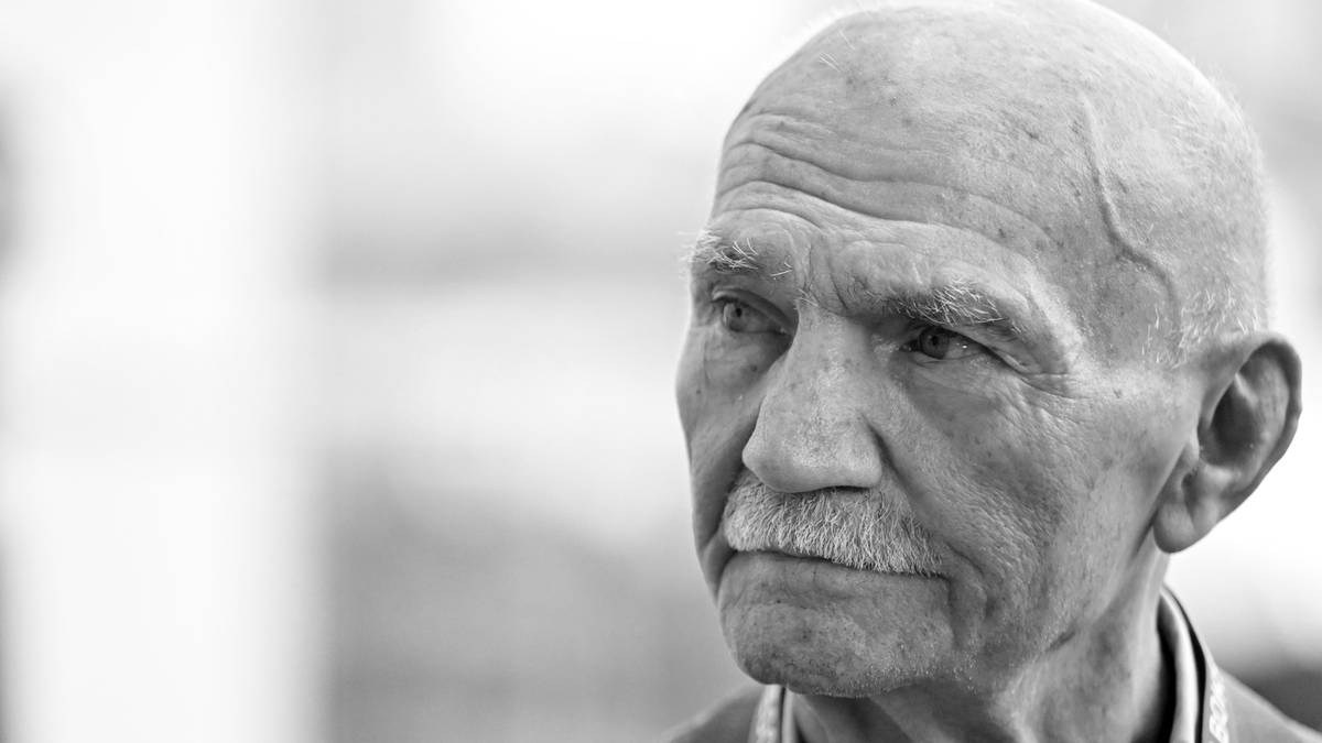 Zbliżenie na twarz starszego mężczyzny z siwymi włosami i wąsami, ubranego w garnitur i koszulę, z widoczną szarfą.