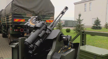 Widok z bliska na zamontowane podw&oacute;jne działko automatyczne, z widocznym magazynkiem z amunicją. W tle znajduje się wojskowy pojazd ciężarowy i budynki przemysłowe.