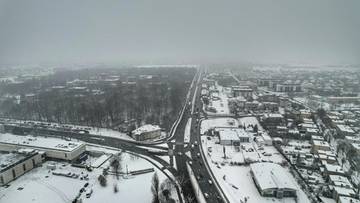 Widok z lotu ptaka na zimowe miasto z drogą prowadzącą przez nie, otoczone drzewami i budynkami pokrytymi śniegiem.