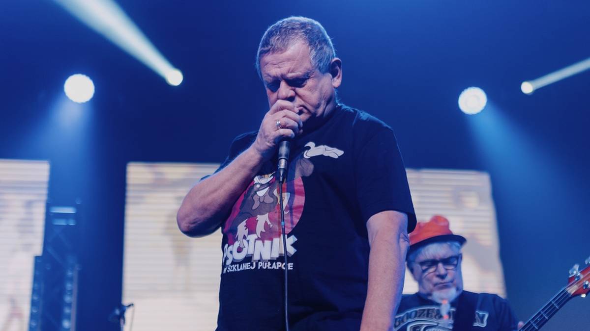 Kazimierz Staszewski, lider zespołu Kult, śpiewa do mikrofonu na scenie podczas koncertu, ubrany w czarną koszulkę i spodnie. Obok niego stoją inni muzycy zespołu.