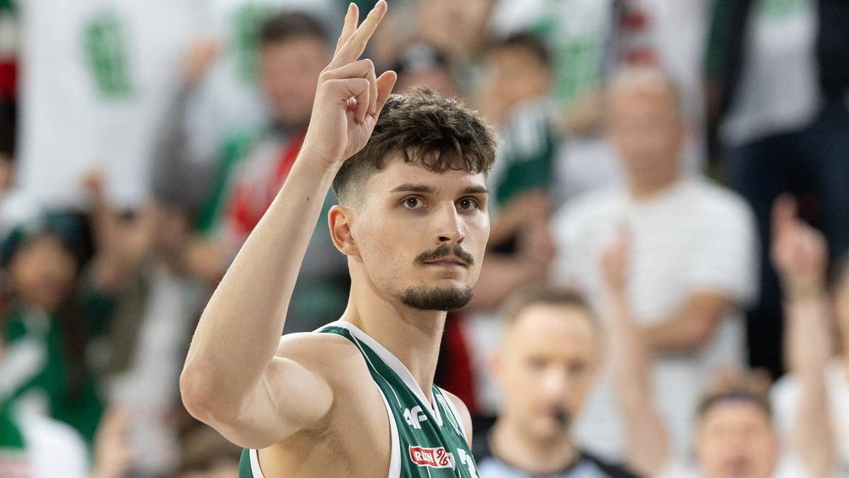 Koszykarz Legii Warszawa w zielonym stroju unosi palec wskazujący do g&oacute;ry na tle trybun z kibicami.