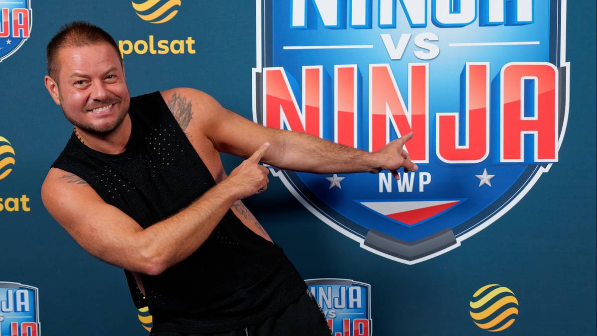 Mężczyzna w czarnym stroju sportowym, stojący przed niebieskim tłem z logo "Ninja vs Ninja", wykonuje taneczny ruch ręką.