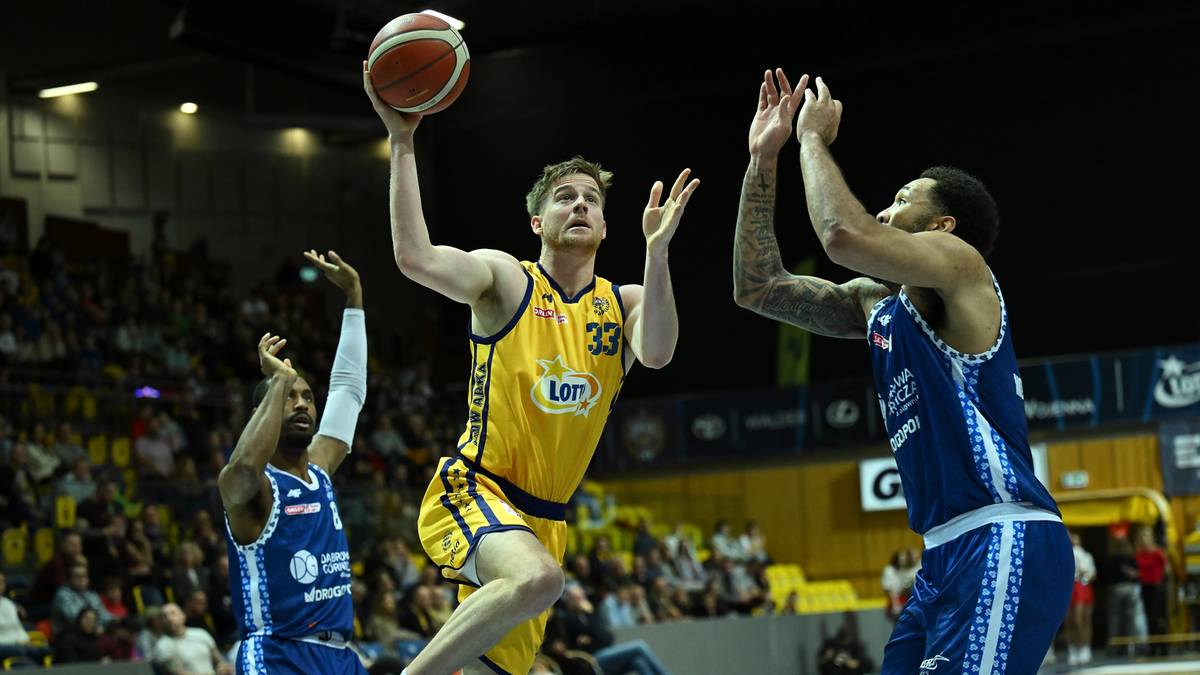 Trzech koszykarzy podczas meczu. Jeden gracz w żółtym stroju z numerem 33 na plecach wykonuje rzut do kosza, podczas gdy dwaj przeciwnicy w niebieskich strojach próbują go zablokować.