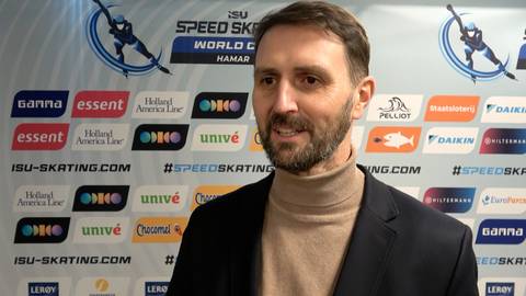 Mężczyzna w średnim wieku z zarostem, ubrany w ciemną marynarkę i beżowy golf, stoi na tle ściany z logotypami i napisami związanymi z łyżwiarstwem szybkim.