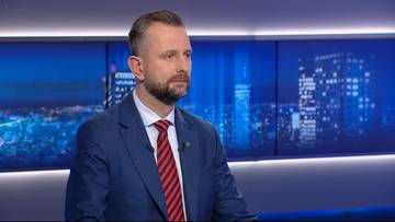 Mężczyzna w garniturze z krawatem siedzi w studiu telewizyjnym na tle panoramy nocnego miasta.