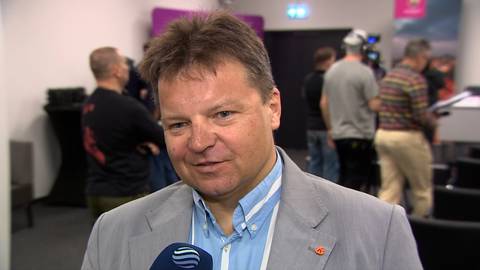 Mężczyzna w szarym garniturze i niebieskiej koszuli, z identyfikatorami na piersi, udziela wywiadu. W tle widoczne są inne osoby w rozmytej scenerii.