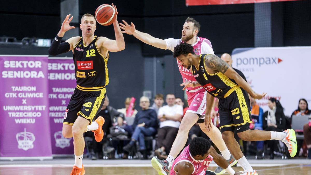 Dw&oacute;ch zawodnik&oacute;w w czarno-ż&oacute;łtych strojach koszykarskich walczy o piłkę z przeciwnikiem w r&oacute;żowym stroju, kt&oacute;ry upadł na parkiet.