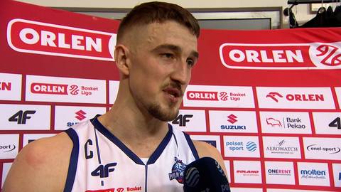 Tomasz Gielo, koszykarz w białej koszulce z logo 4F, udziela wywiadu przed tablicami sponsorskimi z logo ORLEN.