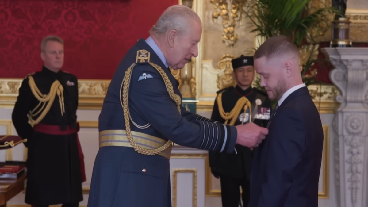 Król Karol III w mundurze lotniczym wręcza Medal Jerzego mężczyźnie w garniturze, podczas uroczystości w sali reprezentacyjnej.