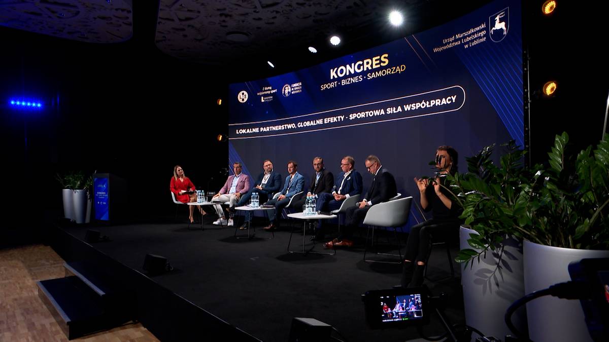 Grupa ośmiu osób siedzi na scenie podczas panelu dyskusyjnego. Przed nimi stoliki, a w tle duży ekran z logotypem i napisem "Kongres Sport, Biznes, Samorząd".
