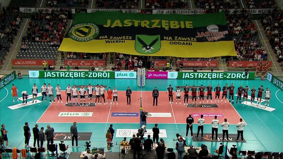 Drużyny siatkarskie ustawione na boisku przed meczem, na trybunach widoczni kibice, za boiskiem duża zielona flaga z napisem "JASTRZĘBIE".