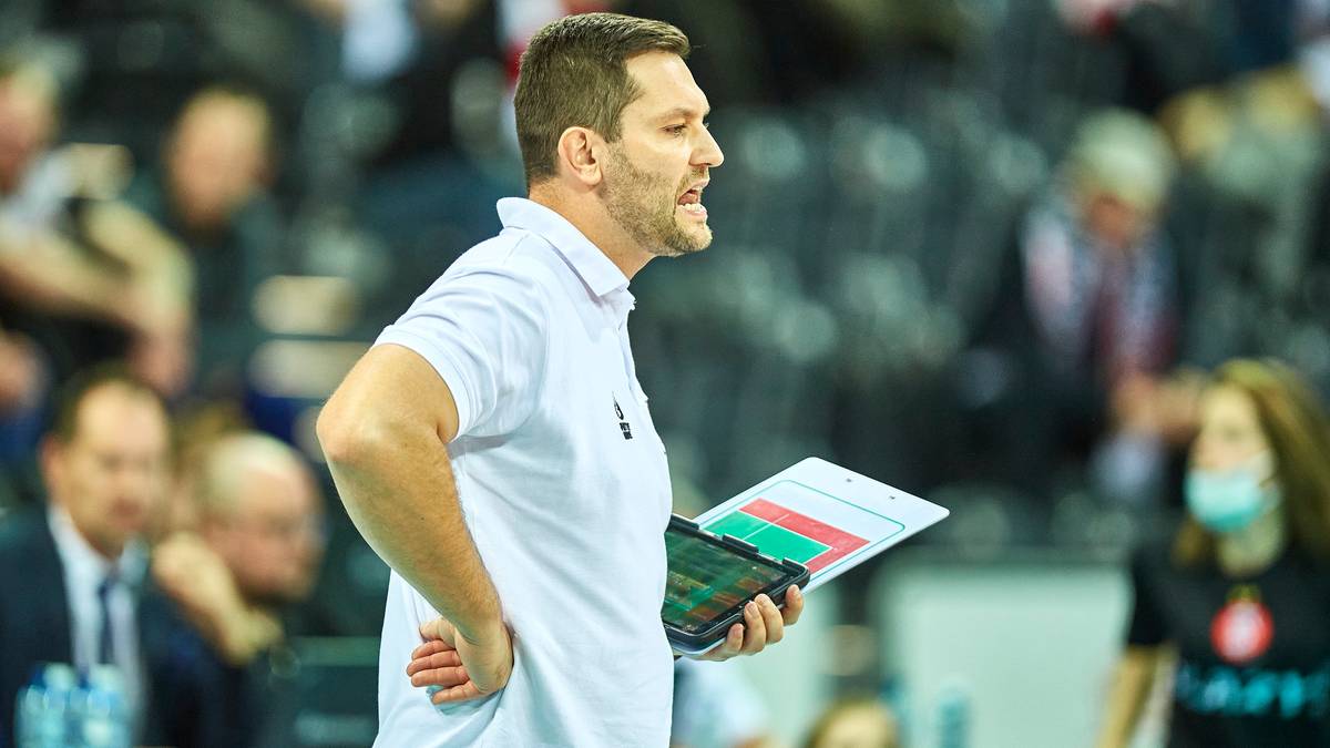 Trener siatkówki z planszą taktyczną w ręku, z wyraźnym wyrazem twarzy, stoi na tle publiczności na trybunach.