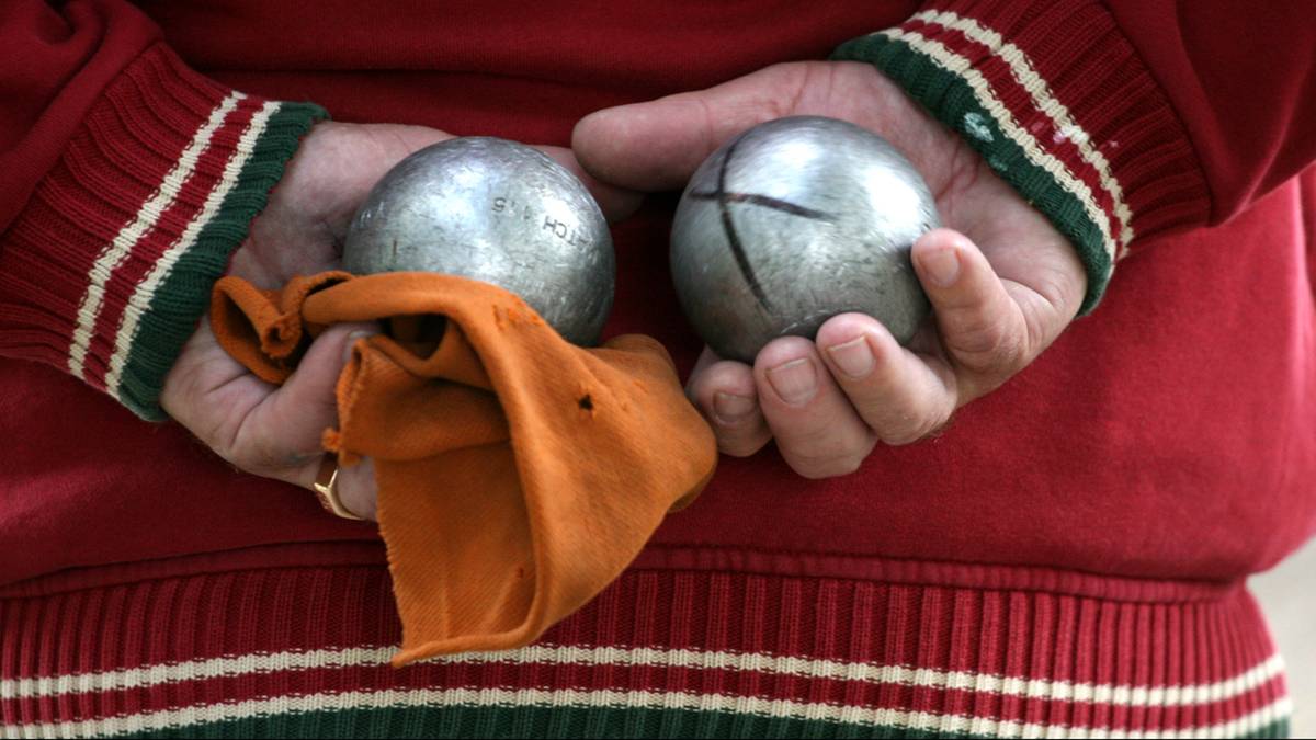 Dwie metalowe kule do gry w petanque w dłoniach osoby ubranej w czerwoną bluzę z zielono-białymi pasami na rękawach. Obok kul znajduje się pomarańczowa chusta.