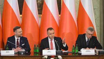 Trzech mężczyzn siedzi przy stole konferencyjnym na tle flag Polski. Jeden z mężczyzn gestykuluje podczas przem&oacute;wienia.