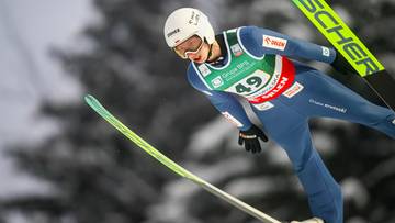 Skoczek narciarski w niebieskim kombinezonie i białym kasku wykonuje skok, z nartami skierowanymi w d&oacute;ł.