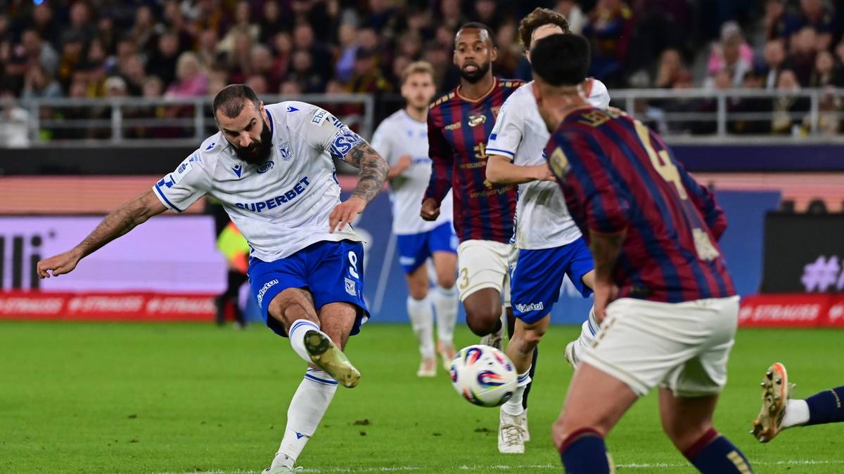 Piłkarz Pogoni Szczecin w białej koszulce i niebieskich spodenkach oddaje strzał na bramkę, podczas gdy zawodnik Lecha Poznań w granatowo-czerwonym stroju pr&oacute;buje go zablokować.