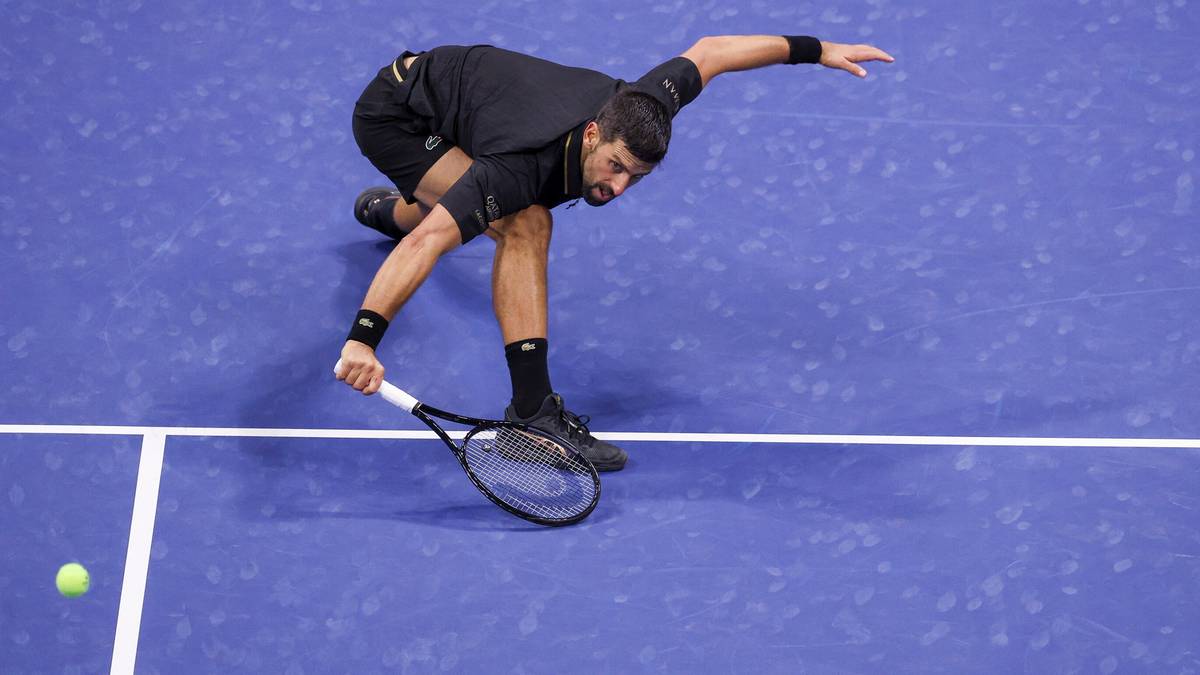 Novak Djokovic wykonuje potężny serwis podczas meczu na US Open. Jest w ruchu, z rakietą w dłoni, gotowy do uderzenia piłki, która jest widoczna w dolnej części kadru.