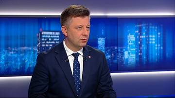 Michał Dworczyk w garniturze i krawacie w studio telewizyjnym na tle z motywem miejskim.
