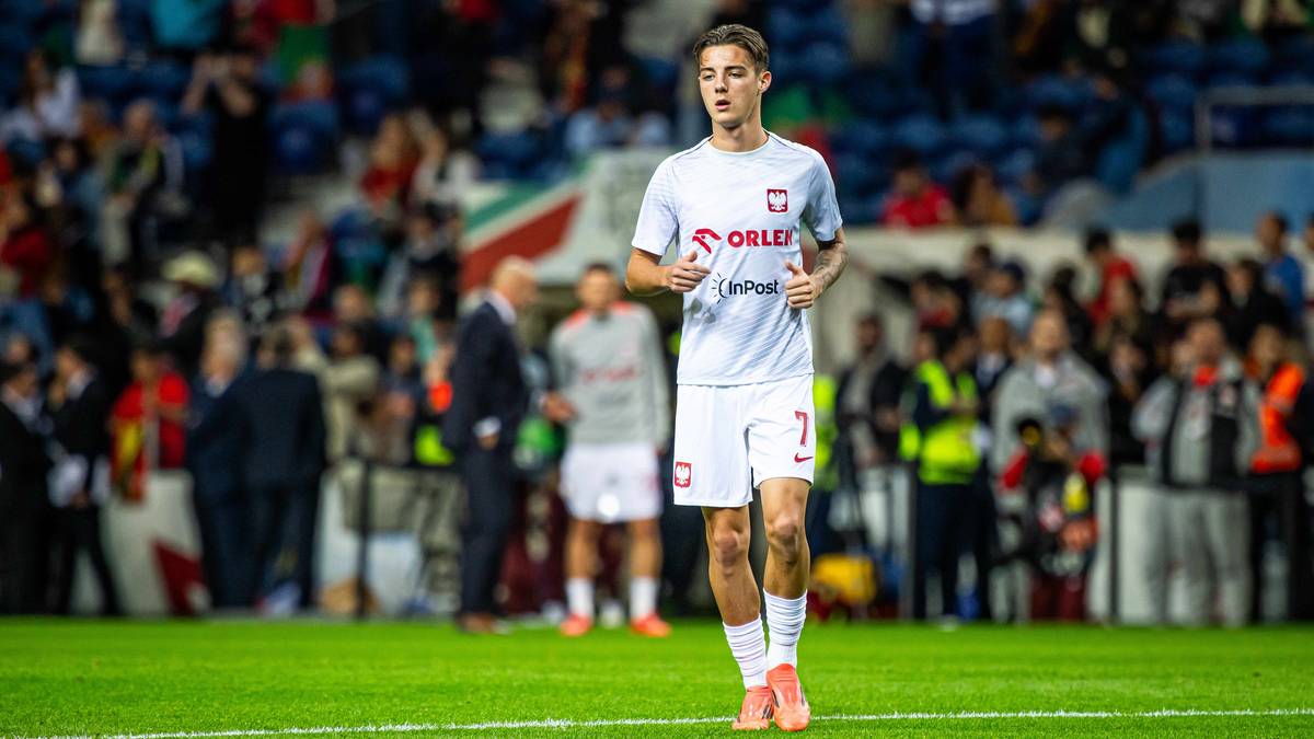 Młody piłkarz w białej koszulce reprezentacji Polski biegnie po boisku podczas meczu.