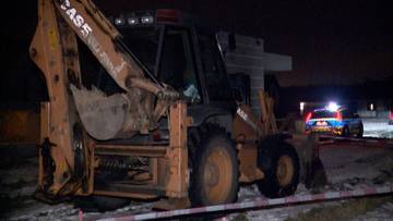 Koparko-ładowarka na placu budowy w nocy, w tle policyjne światła.